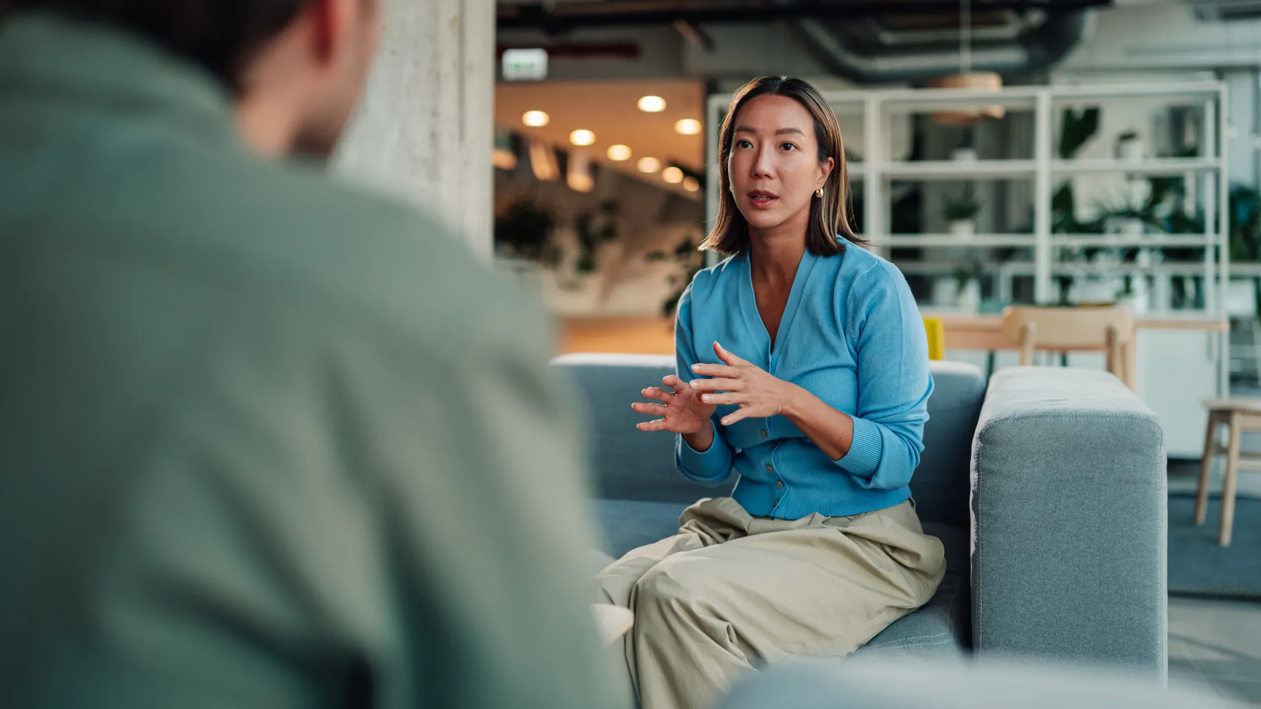 A woman in a blue cardigan in conversation with a colleague in an office lounge