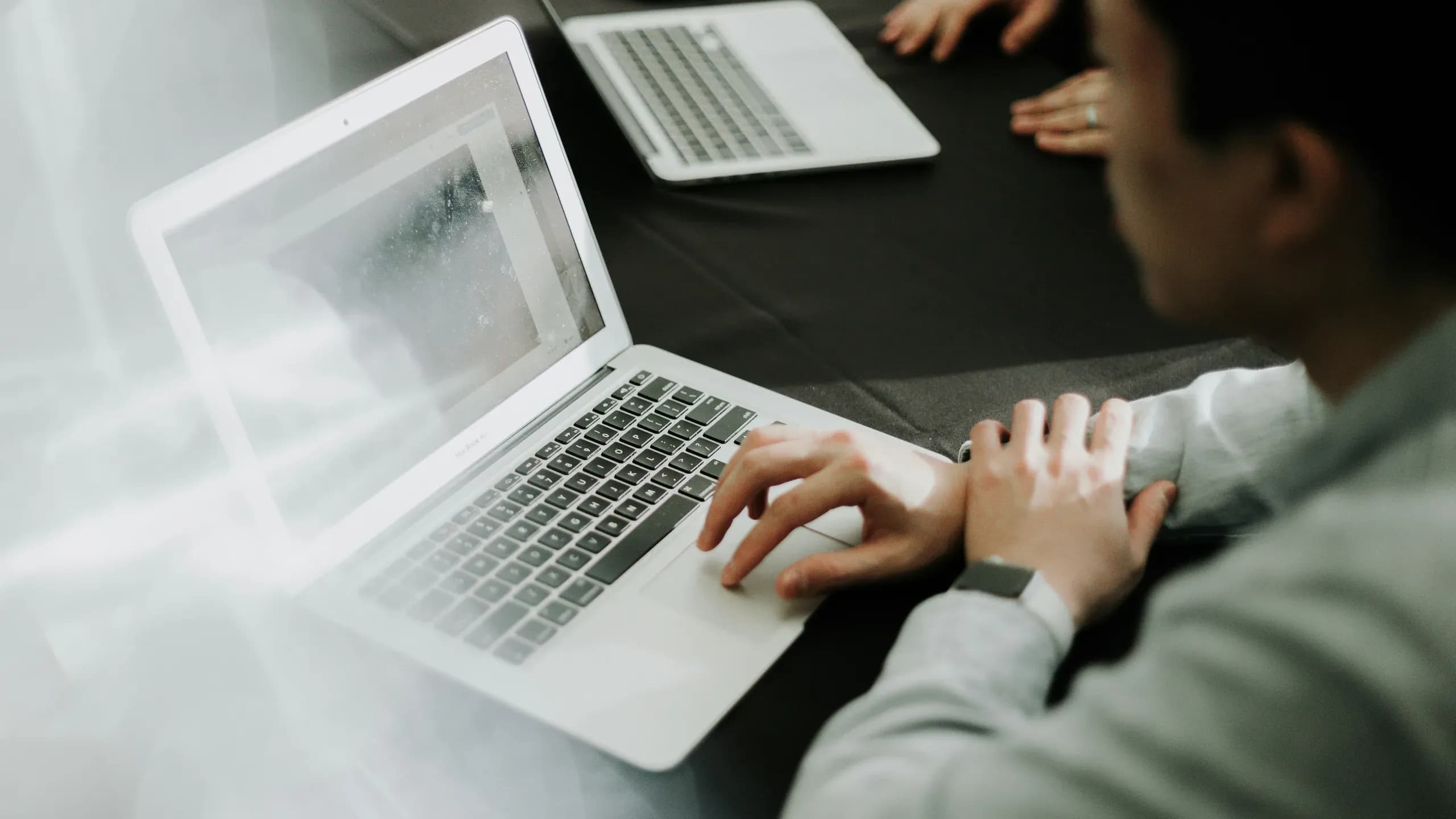 A person typing on a laptop at a dark table, with a second laptop and a colleague’s hands across from them