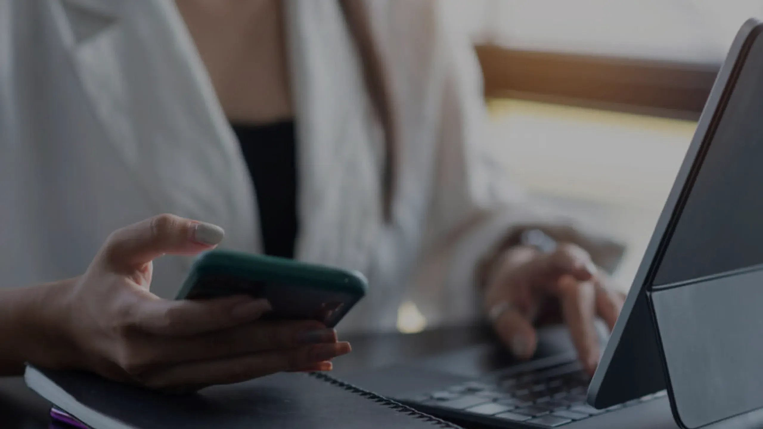 A person at a desk holding a phone in one hand and typing on a laptop with the other
