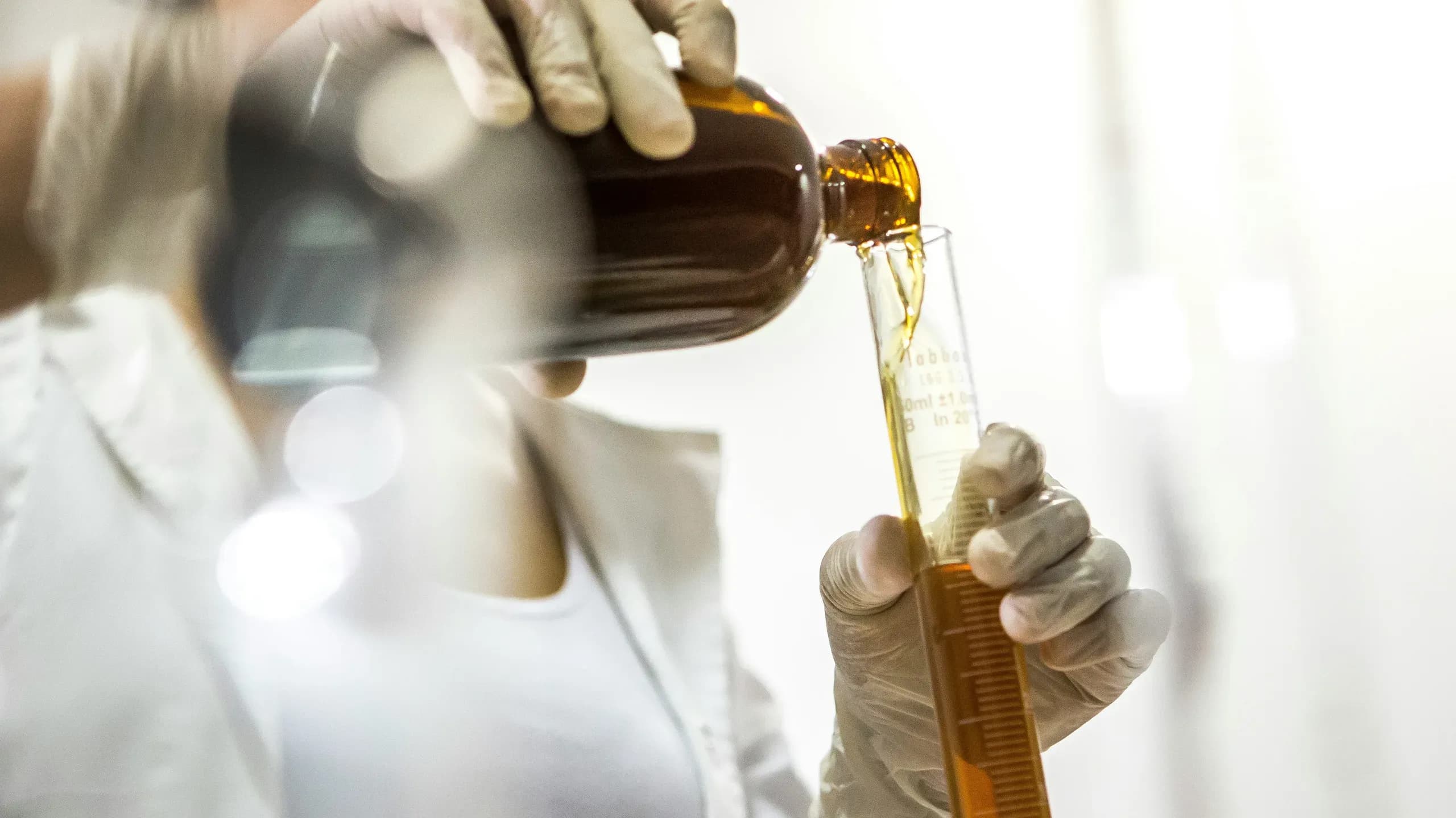 A lab worker in white gloves pouring an amber liquid from a bottle into a graduated cylinder