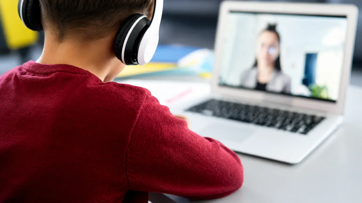 A child in a red sweater and headphones at a laptop, with a teacher on screen in a video lesson