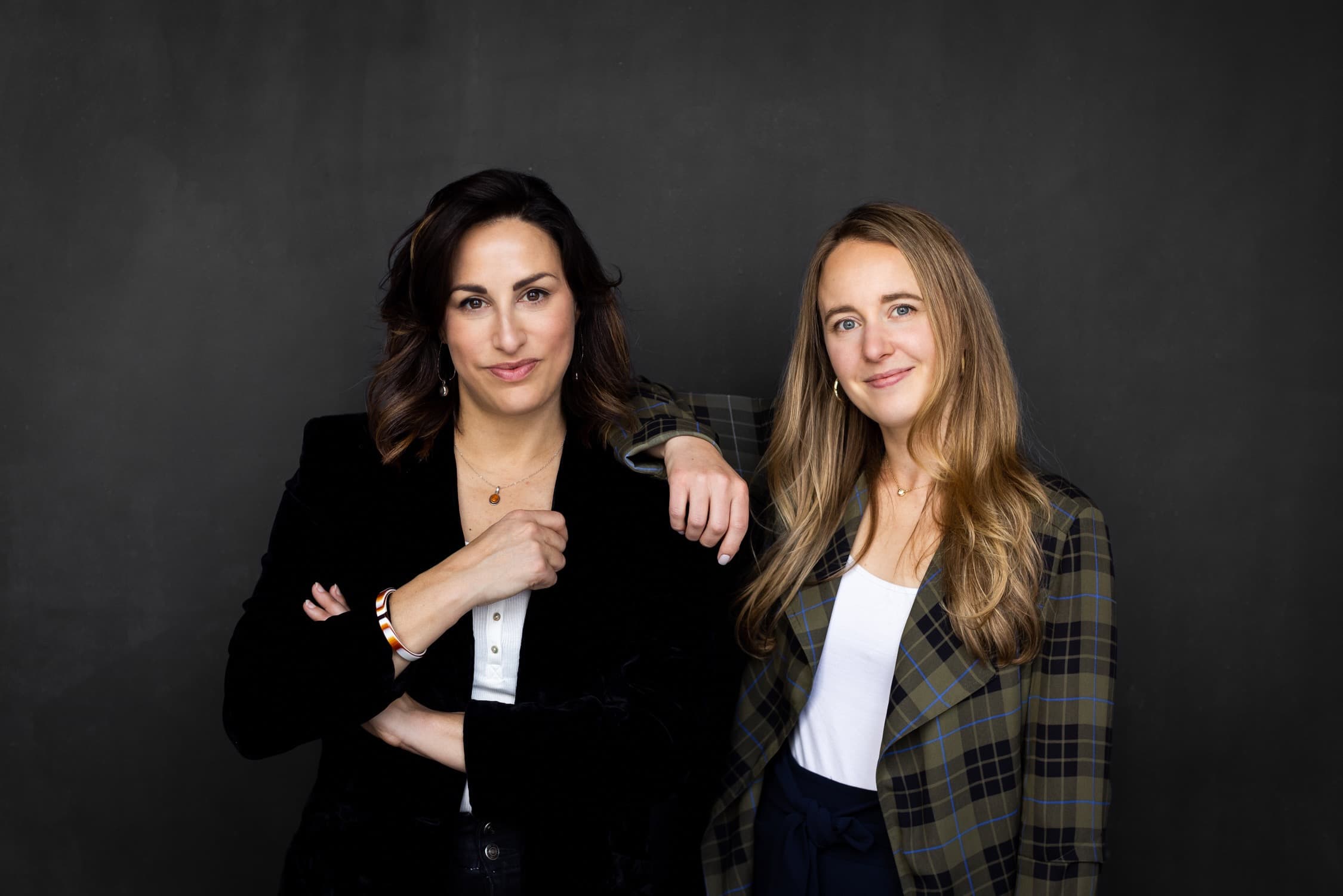Two women stand side by side against a dark grey backdrop, one resting her arm on the other’s shoulder.