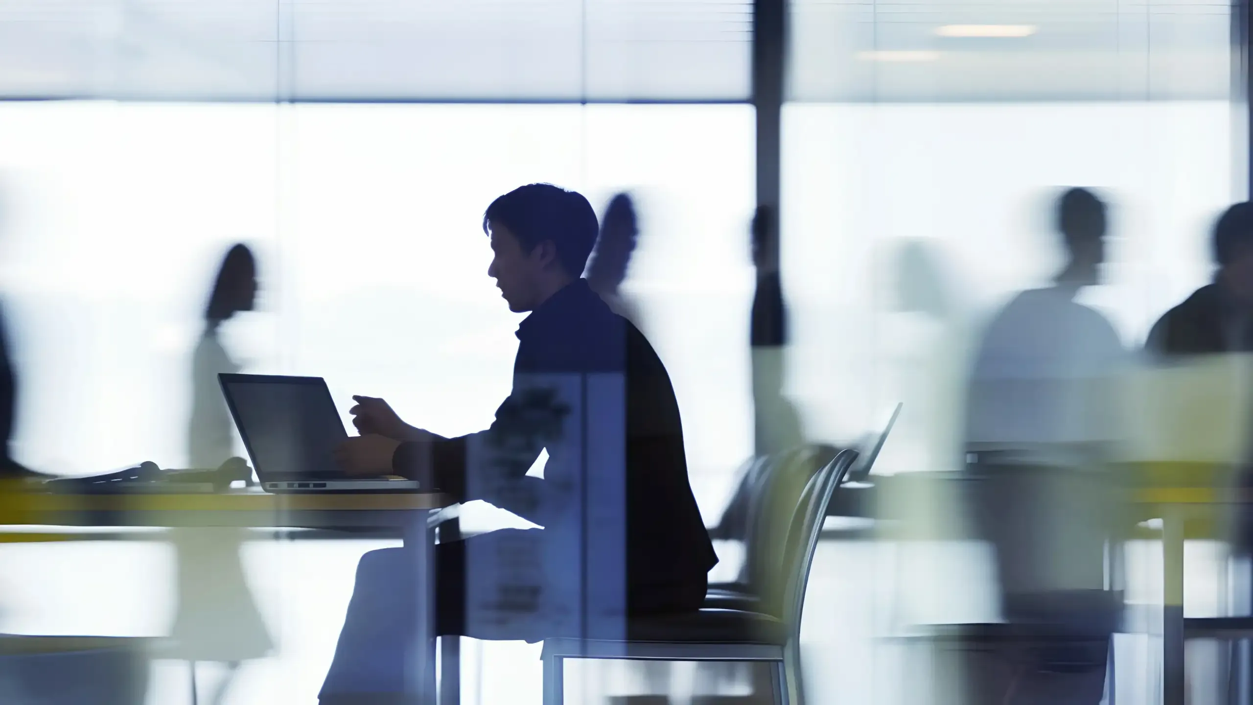 A person working at a laptop in a glass-walled office, colleagues passing behind
