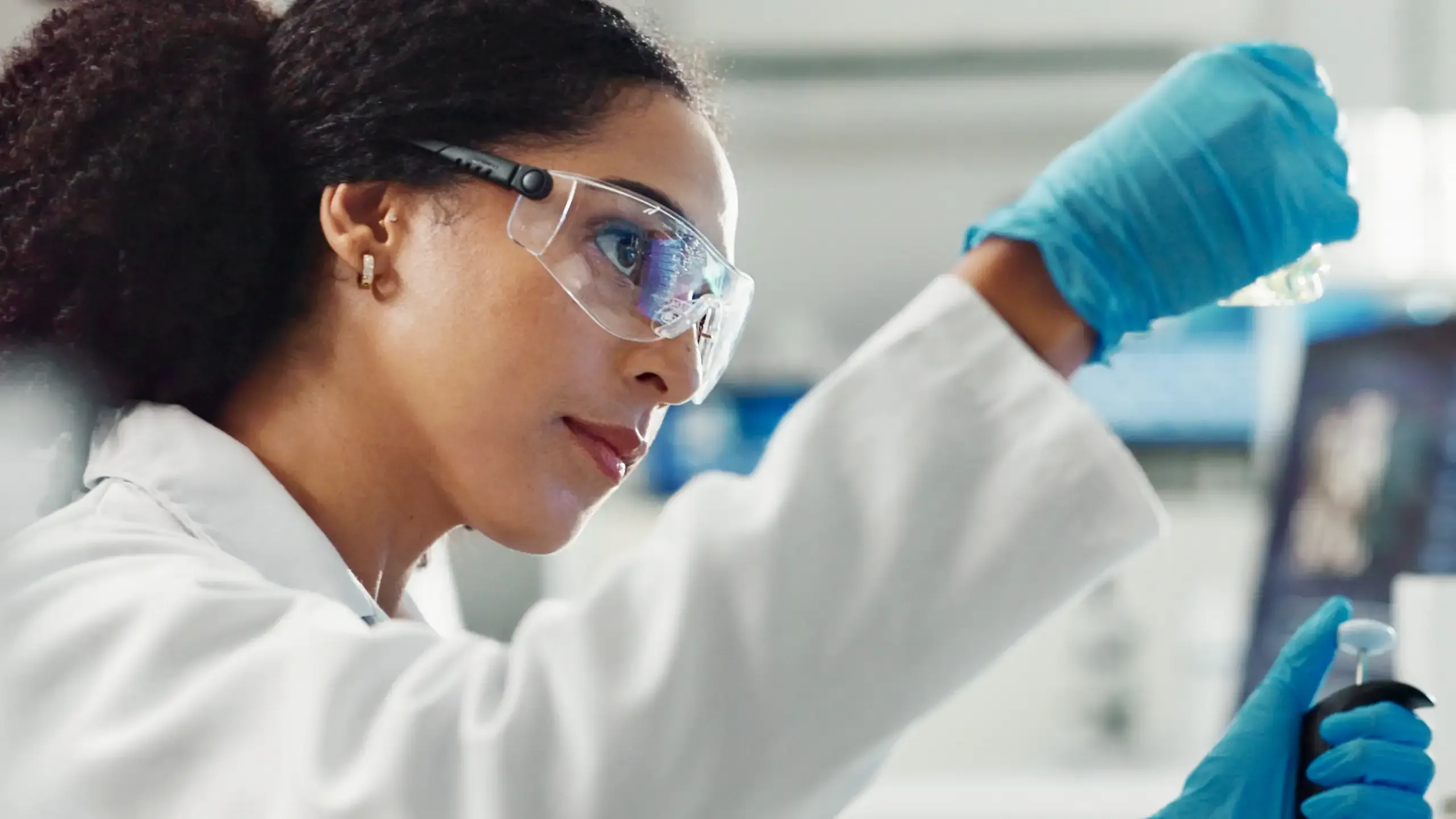 A scientist in safety glasses and blue gloves examining a sample in a laboratory