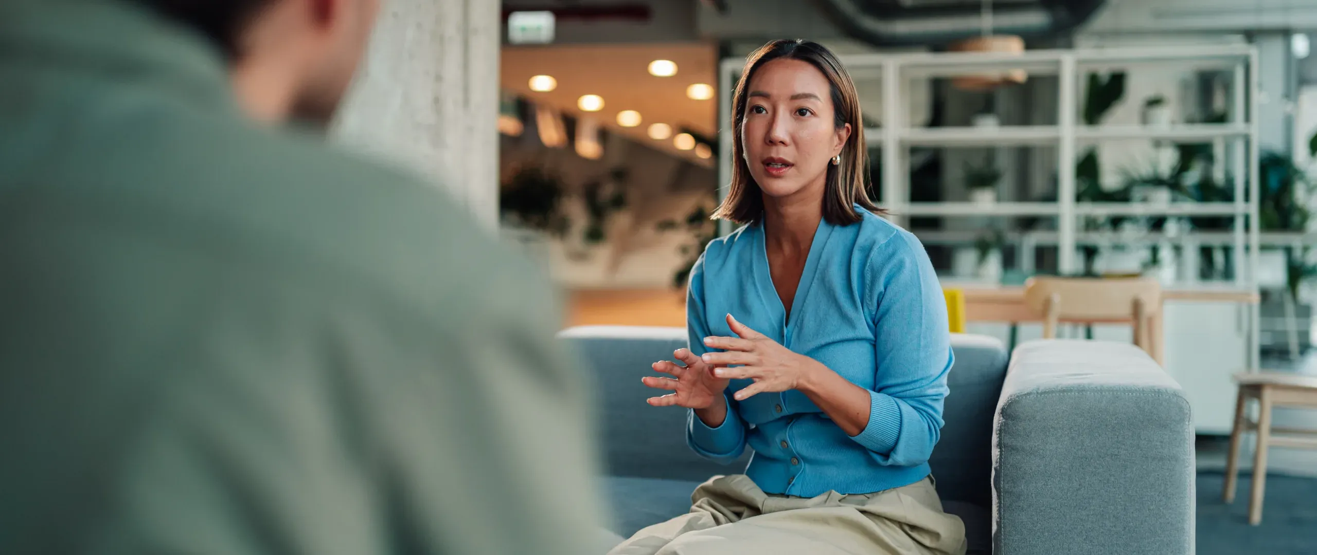 A woman in a blue cardigan in conversation with a colleague in an office lounge