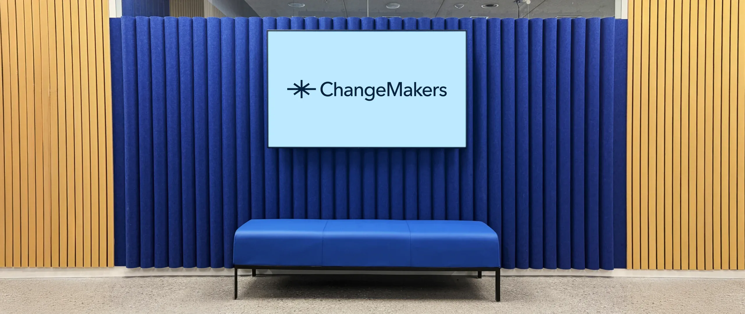 A ChangeMakers office lounge: a blue bench in front of a wall of deep-blue acoustic panels, with the ChangeMakers logo on a wall-mounted screen and wood-slat walls either side