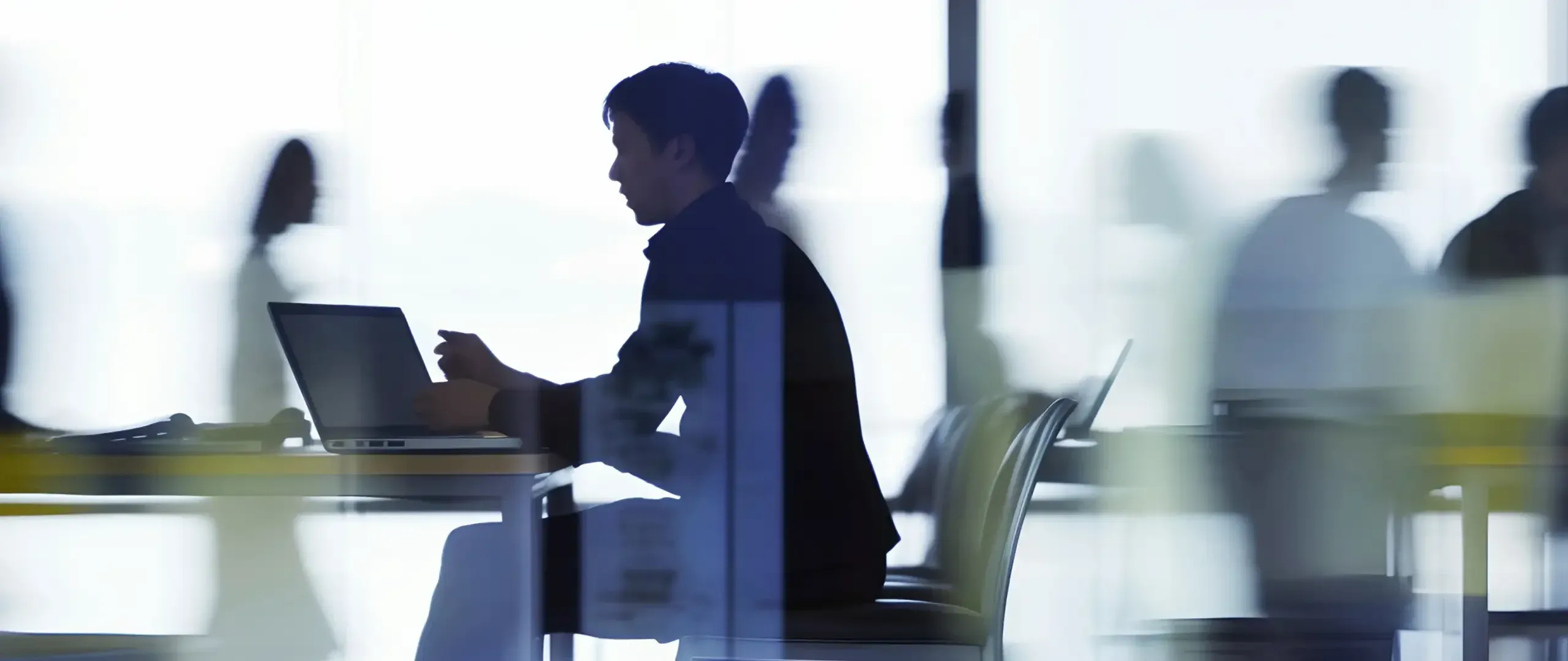 A person working at a laptop at a table, seen in silhouette through glass, with colleagues blurred in motion behind them