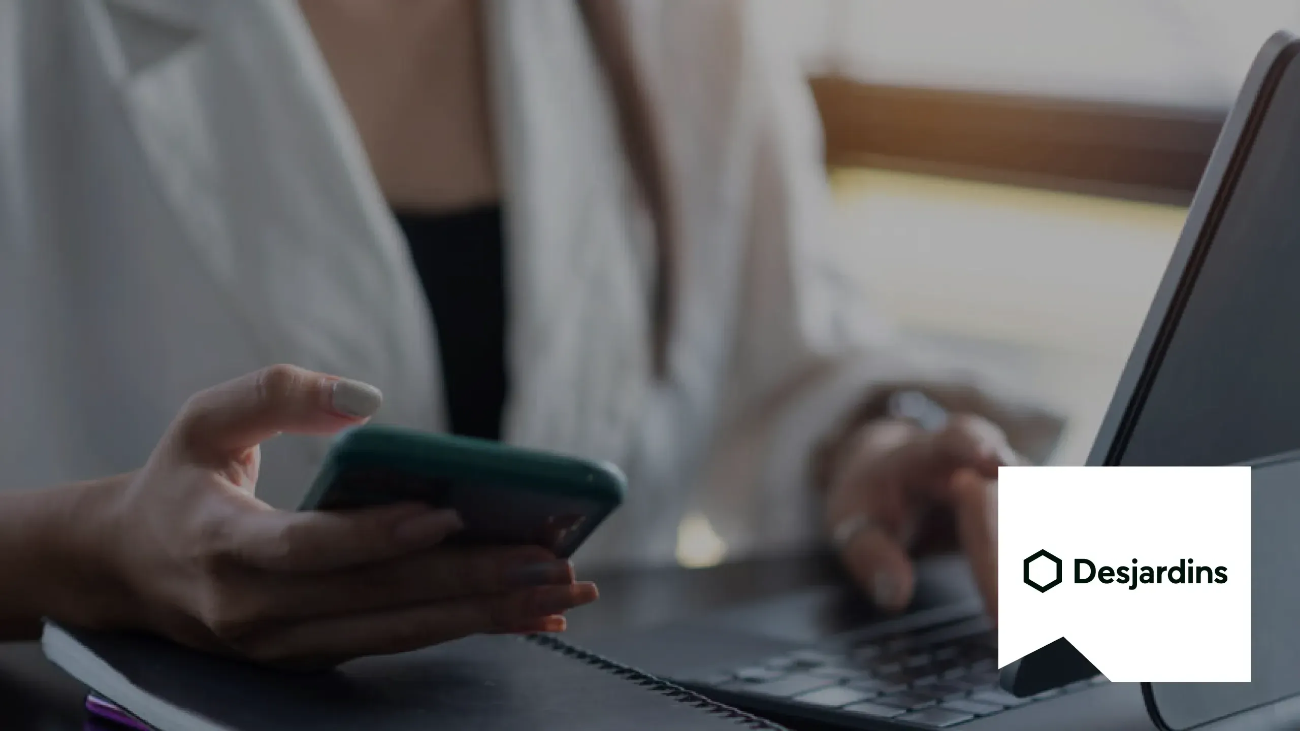 A person at a desk holding a phone in one hand and typing on a laptop with the other, with the Desjardins logo on a white panel in the corner