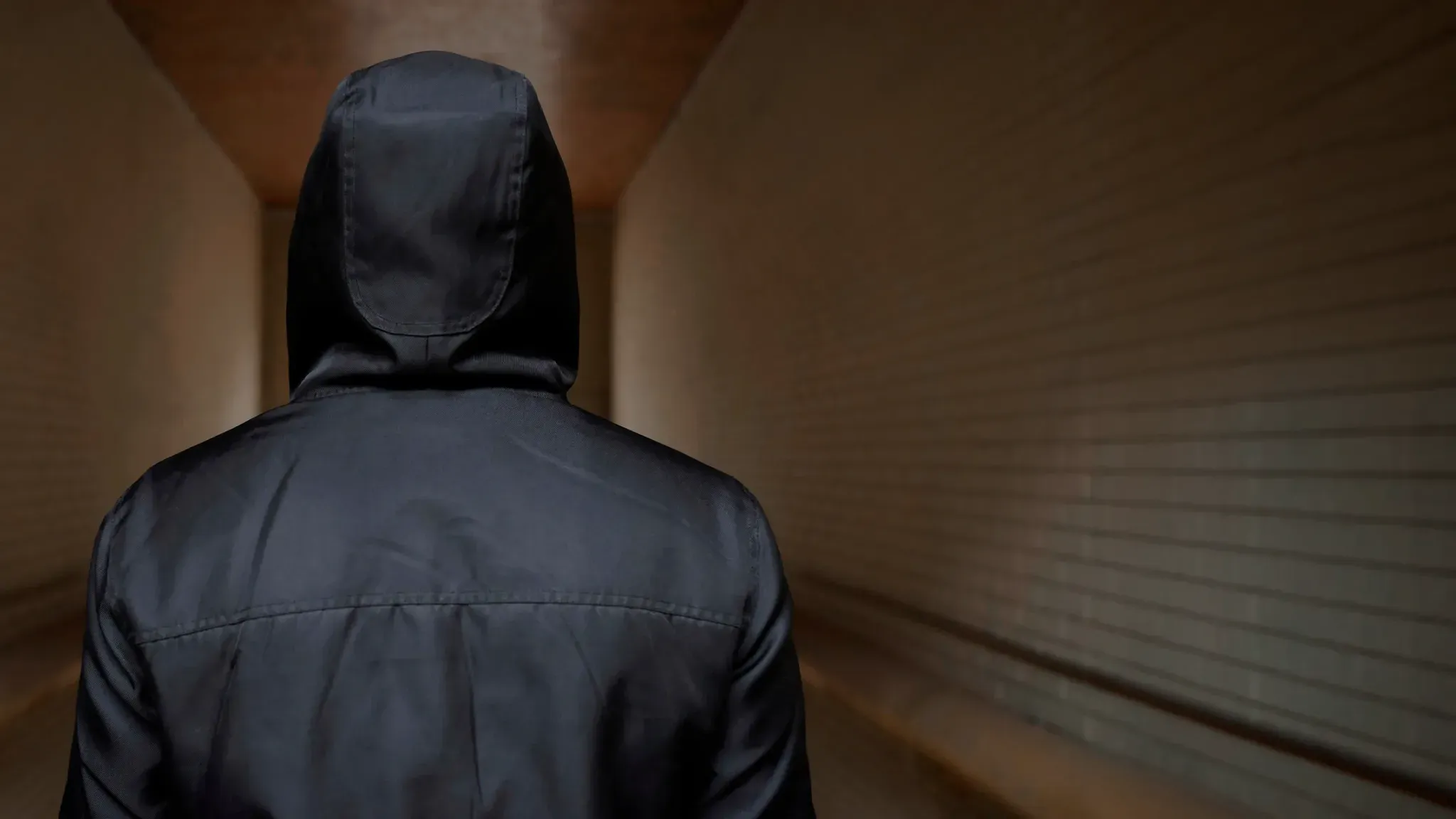 A person in a dark hooded jacket seen from behind in a dim corridor.