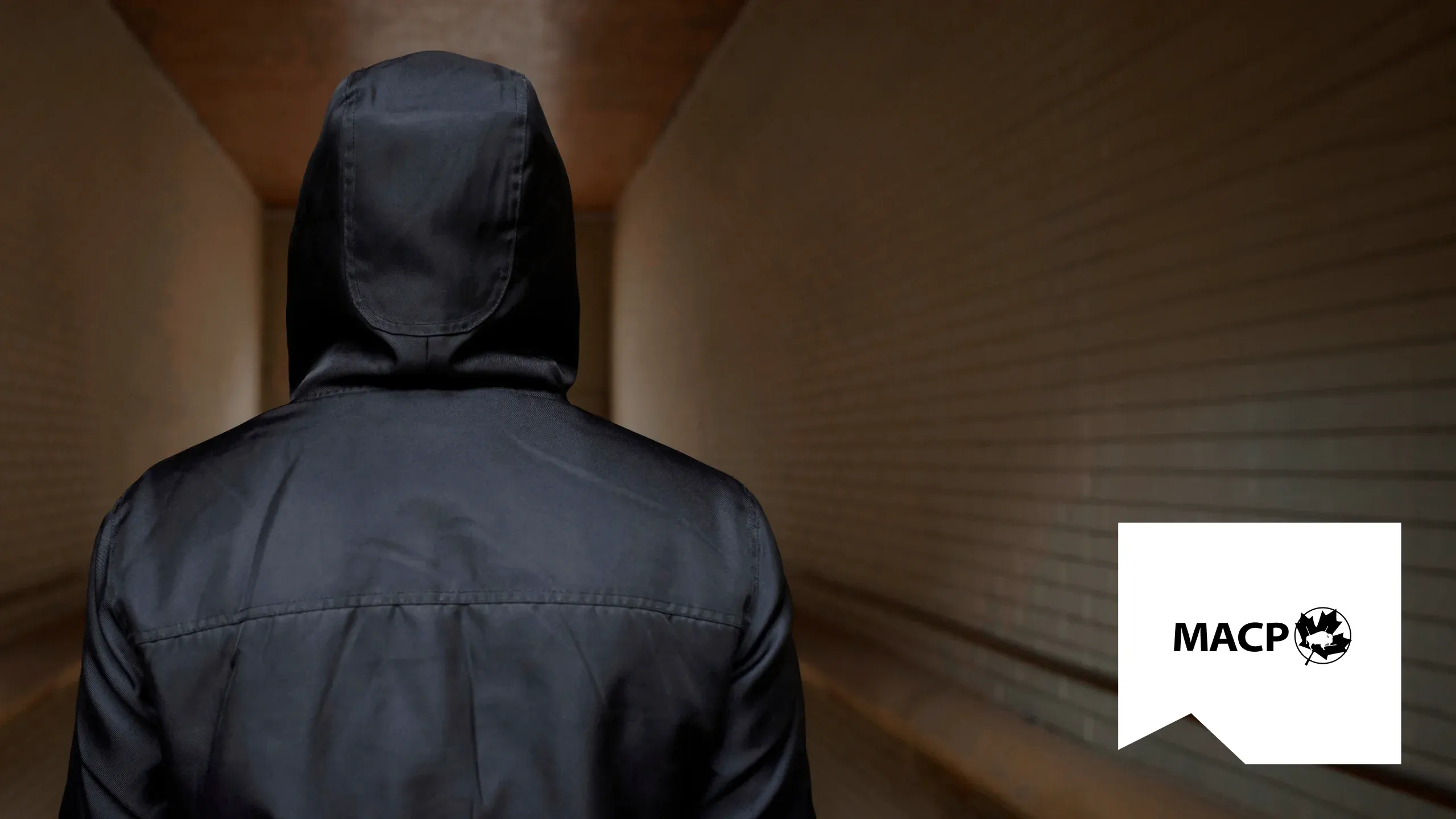 A person in a dark hooded jacket seen from behind in a dim corridor, with the MACP logo on a white panel in the corner